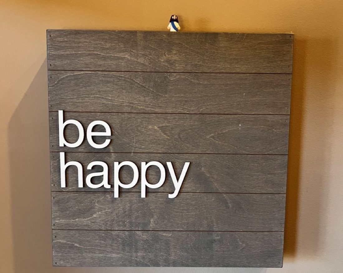 A Mini Jesus figure perched on top of a wooden wall sign that reads be happy