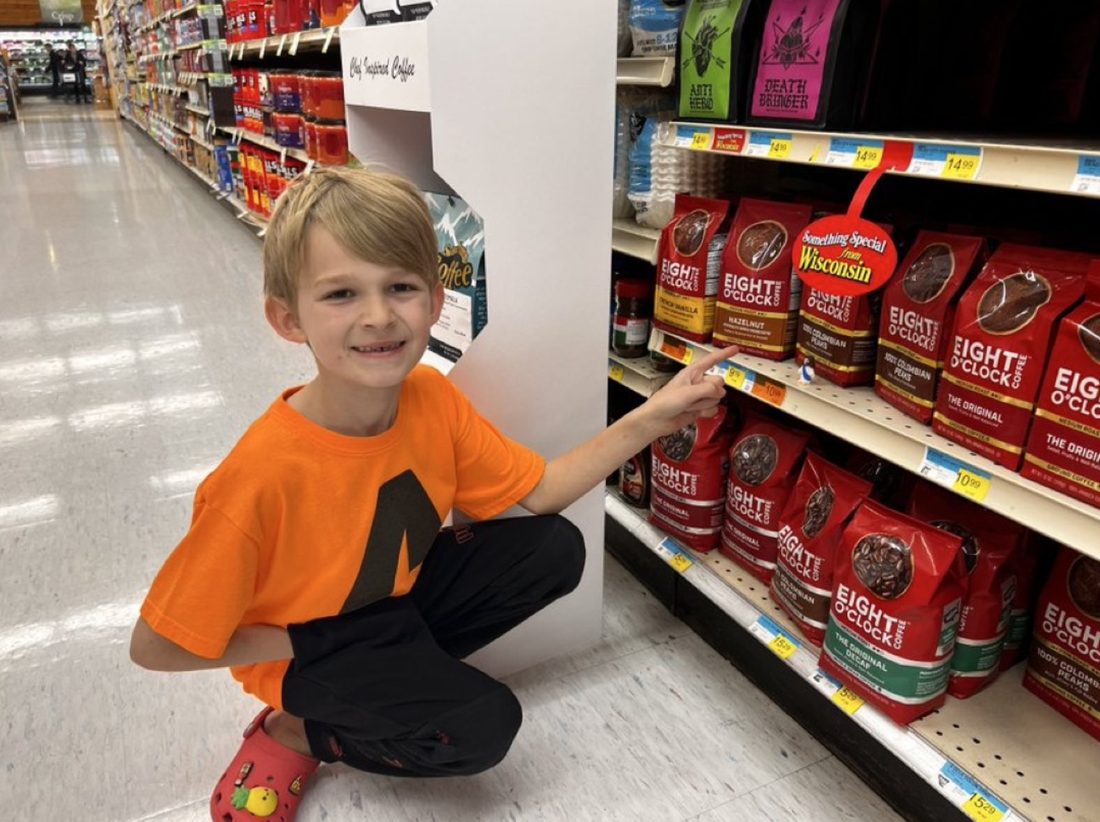 A young boy kneeling in a grocery store aisle, smiling, next to a Mini Jesus figure tucked on the shelf