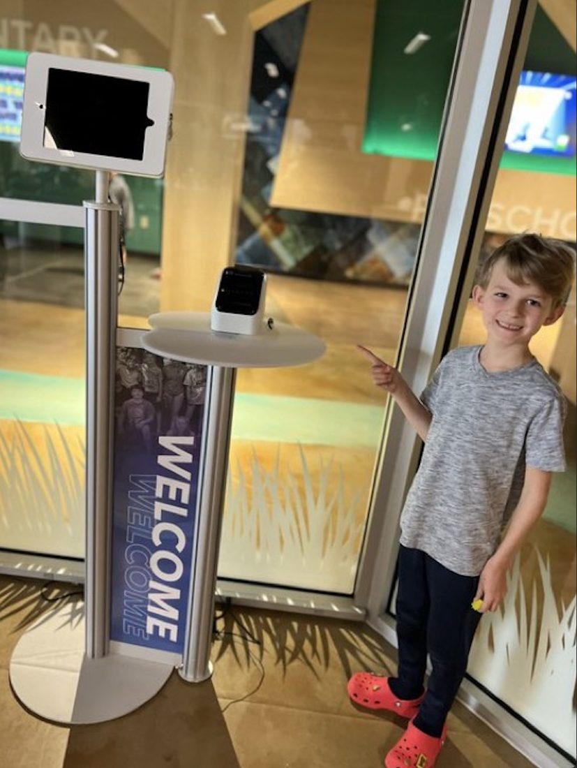 A boy pointing at a Mini Jesus figure left by a welcome sign in a lobby